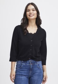 Svart cardigan med V-hals, 6 knappar fram, och trekvartsärmar. Buren över en spets camisole, ihop med blå jeans.