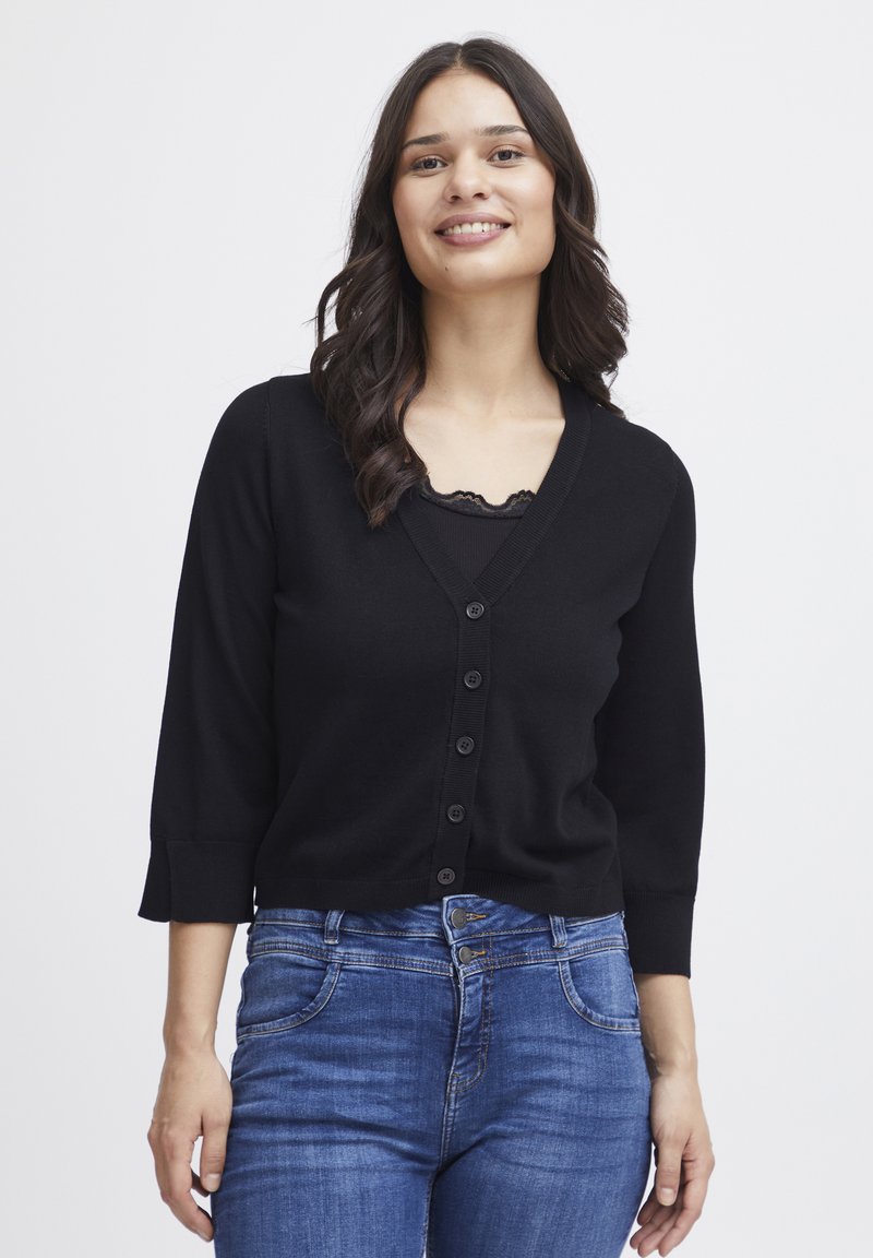 Schwarze Strickjacke mit V-Ausschnitt, 6 Knöpfen vorne und Dreiviertelärmeln. Getragen über einem Spitzen-Cami, kombiniert mit blauen Jeans.