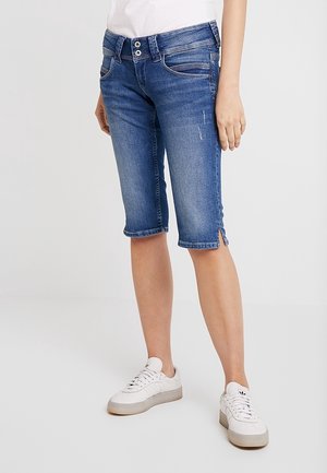 Persoon die blauw denim shorts van middellange lengte draagt met zijsplitten, witte sneakers en een wit shirt, staand tegen een effen achtergrond.