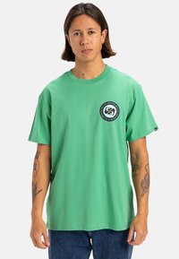 Camiseta de algodón verde con un logo redondo en el pecho izquierdo. Mangas cortas, corte relajado, costuras visibles y etiqueta en el lado inferior.