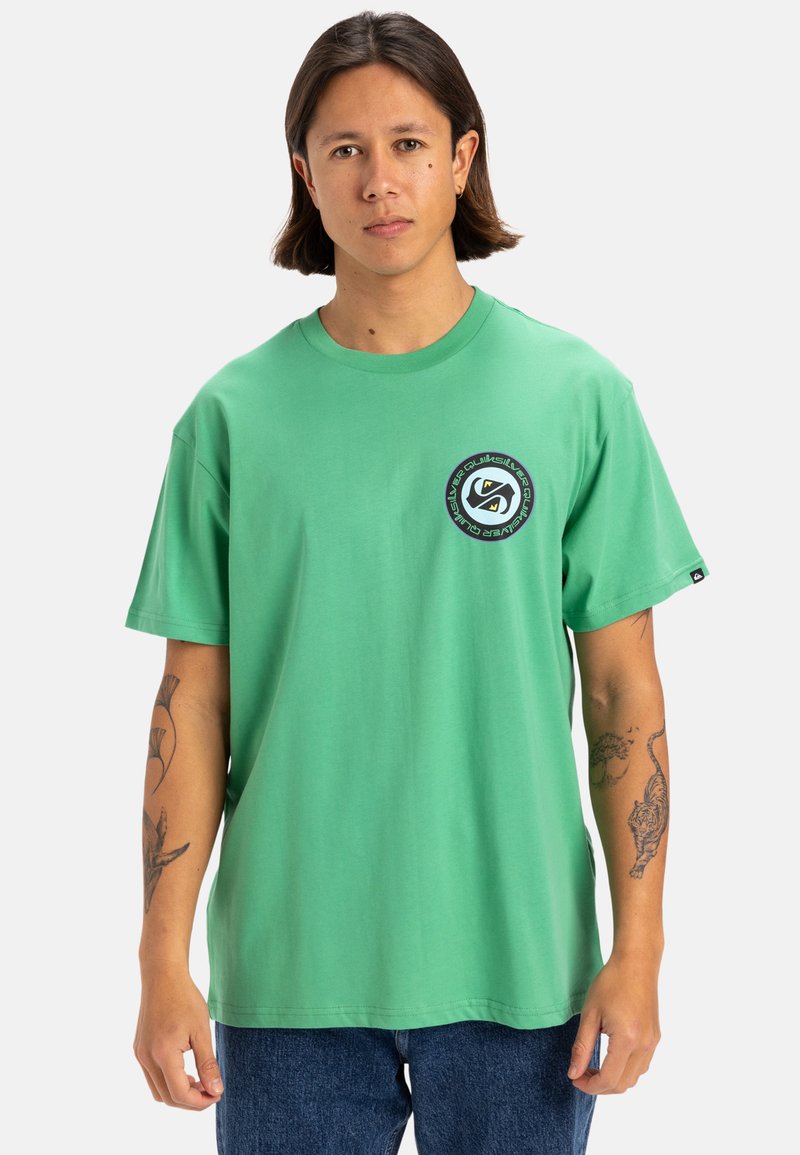 Camiseta de algodón verde con un logo redondo en el pecho izquierdo. Mangas cortas, corte relajado, costuras visibles y etiqueta en el lado inferior.