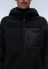 Chaqueta de forro polar negra con una superficie texturizada y esponjosa y un bolsillo en el pecho con cremallera. Presenta una capucha y paneles de acento negros.