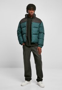 Teal dunjakke med mønstret front, svarte skuldre og høy krage. Kombinert med mørke jeans og hvite sneakers.