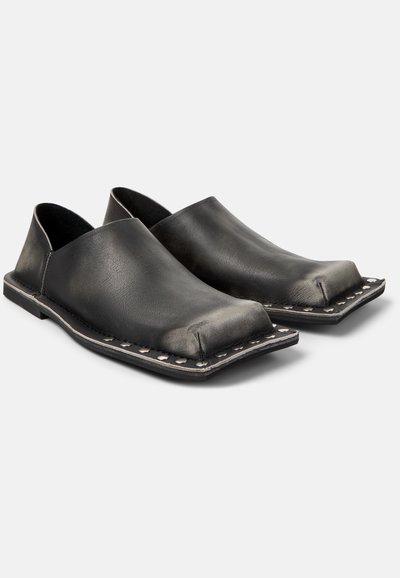 Chaussures plates en cuir noir à enfiler, avec bouts carrés, coutures apparentes et clous métalliques autour des bords de la semelle sur fond blanc.