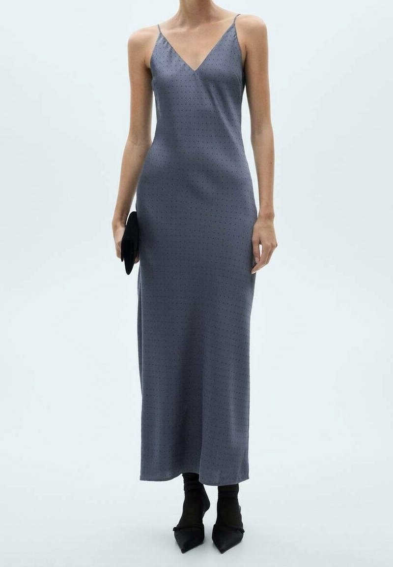 Kobieta w bezrękawną, szaro-anatomię sukienkę do kostek z cienkimi ramiączkami, czarnych, spiczastych butach oraz trzymająca czarną kopertówkę.