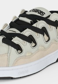 Κοντινή λήψη ενός μπεζ και άσπρου sneaker Osiris με μαύρα κορδόνια και ορατό λογότυπο στη γλώσσα και στη σόλα.