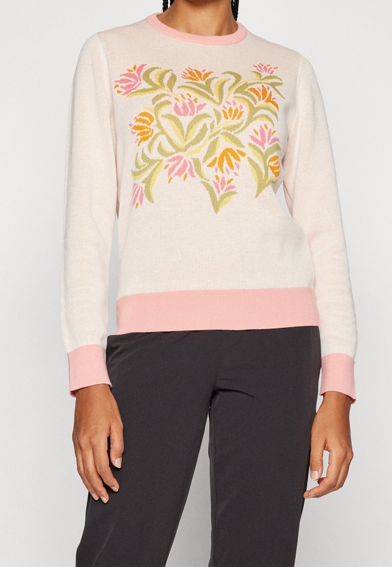 Femme portant un pull rose clair avec un motif floral vert et orange, des poignets et un ourlet roses, et un pantalon noir.