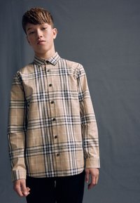 Beiges kariertes Hemd aus Baumwolle, mit schwarz-weißen Karomustern und dunklen Knöpfen. Lange Ärmel und Kragen.