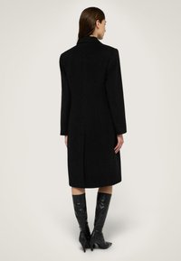 Černý vlněný kabát se strukturovanou siluetou, jednoproudým designem a dlouhými rukávy. Disponuje rovnými zády s centrálním švem.
