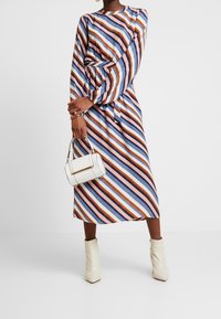 Robe midi rayée en bleu, orange et blanc. Manches longues avec poignets froncés, taille cintrée, associée à un petit sac à main blanc.