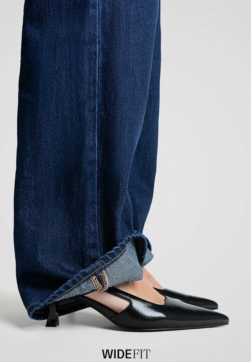 Personne portant un jean large bleu foncé associé à des chaussures noires à bout pointu et à talon bas sur un fond clair.