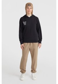 Black hoodie with text "Women of the Wave," paired with beige sweatpants. White sneakers and a tan beanie complete the casual outfit.