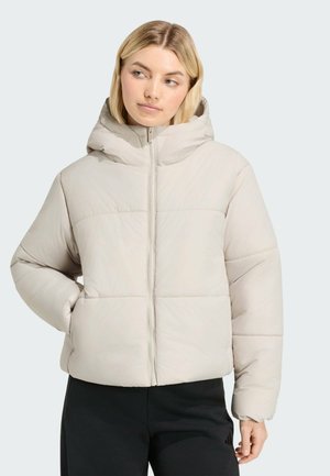 Vrouw draagt een lichtbeige donzen jas met capuchon, dichtgeritst, en zwarte broek, staand met één hand in de zak.