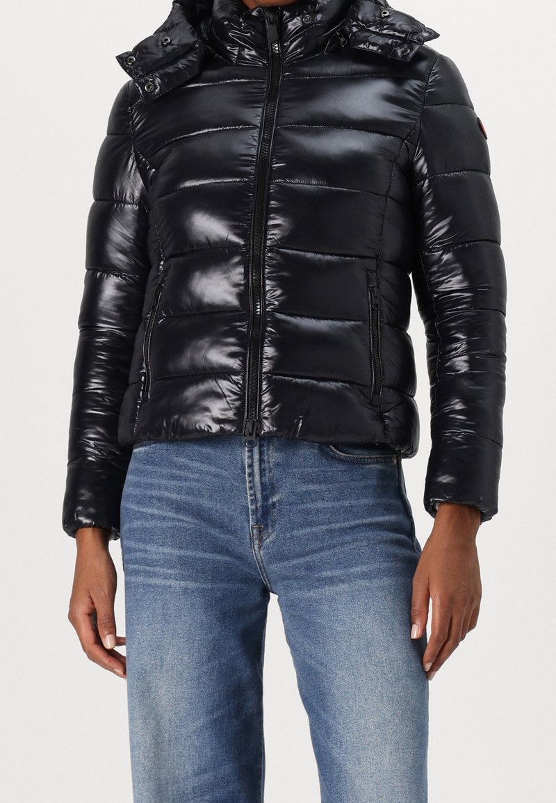 Veste matelassée noire brillante avec fermeture éclair frontale et deux poches zippées, portée avec un jean en denim bleu par une personne sur un fond clair uni.