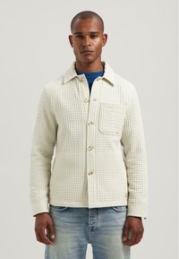 Giacca bianca testurizzata con colletto, tasca frontale e chiusura a bottoni; abbinata a un maglione blu sotto e jeans di denim chiaro.
