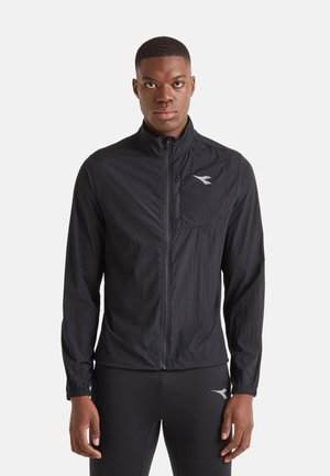Giovane uomo che indossa una giacca sportiva nera con zip e pantaloni sportivi neri con un piccolo logo bianco su entrambi gli indumenti.