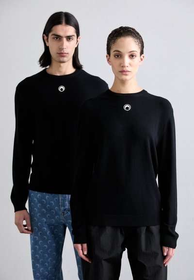 Due persone indossano maglioni neri a maglia con un logo circolare sul petto. Lo sfondo è neutro e la donna indossa pantaloni neri larghi, mentre l'uomo ha jeans blu con un motivo.
