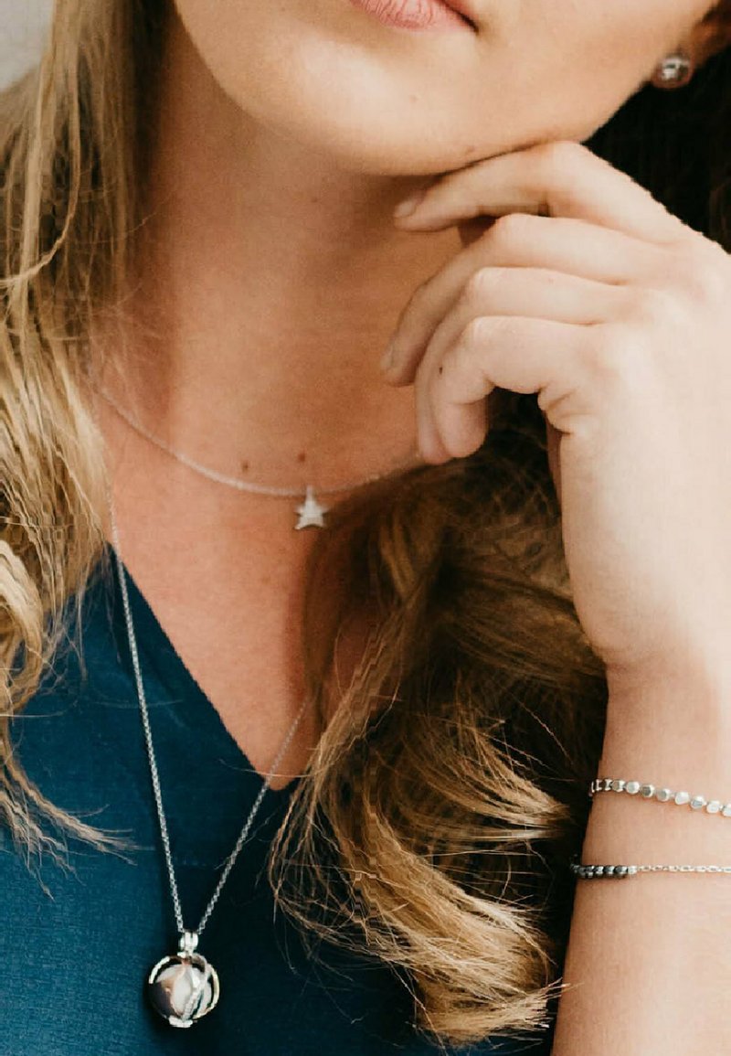 Silverhalsband staplade på en mörk topp, med en stjärnhänge och en rund orb-charm. Handleden visar ett delikat silverarmband med pärlor.