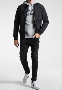 Giacca bomber nera trapuntata indossata sopra una felpa grigia con cappuccio, abbinata a jeans scuri e sneaker bianche con dettagli scuri.