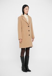 Abrigo de lana beige con cuello de muesca, cinco botones negros y bolsillos laterales, llevado sobre un suéter color crema, combinado con botas altas negras hasta la rodilla.