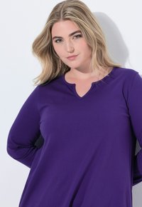 Fioletowa bluzka z długim rękawem i dekoltem w serek, wykonana z gładkiego materiału. Posiada luźny krój i subtelne detale szycia przy kołnierzu.