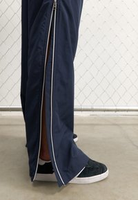 Pantalons de sport navy avec fermetures éclair sur les côtés, fabriqués dans un tissu léger. Dotés d'une fermeture éclair blanche contrastante et de baskets en daim noires avec des semelles blanches.