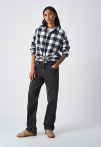 Schwarz-weißer karierten Button-Up-Hemd, das an der Taille gebunden ist, kombiniert mit dunklen Jeans und beigen Sneakers vor einem schlichten Hintergrund.
