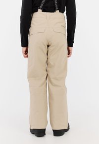 Beige sneeuwbroeken met verstelbare zwarte bandjes, achterzakken, taps toelopende pijpen en een gladde textuur, gedragen met zwarte laarzen.