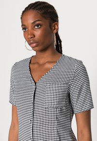 Camisa de gingham preto e branco de mangas curtas com decote em V, dois bolsos no peito e um fecho frontal com botões. Tecido de algodão suave com um padrão clássico.