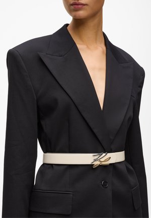 Nő fekete zakóban, széles hajtókákkal, amelyet fehér övvel szorítanak meg a deréknál, az öv fémes, dupla betűs csattal díszített.