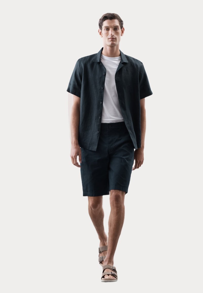 Camisa corta de lino verde oscuro, superpuesta sobre una camiseta blanca y combinada con pantalones cortos azul marino. Usada con sandalias marrones. Diseño suave y casual.