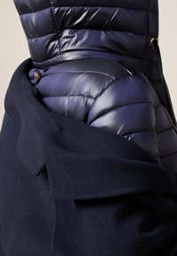 La giacca imbottita presenta un esterno lucido di un blu scuro, trapuntato, e un morbido colletto in lana di un blu navy scuro, con bottoni e dettagli in accentuazione per la chiusura.