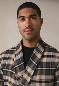 Kariertes Wollbademantel in Beige-, Grau- und Brauntönen, mit einem Schalkragen und sichtbarer Textur, getragen über einem schwarzen Hemd.