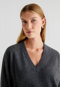 Grijze v-hals trui van zachte stof, met een relaxte pasvorm en geribbelde details bij de hals. Model heeft steil blond haar en lichtblauwe ogen.