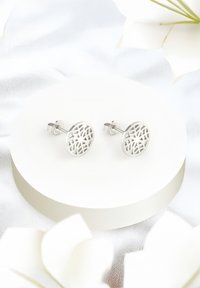 Silvriga studs med ett cirkulärt, utskuret geometriskt mönster. Visade på en rund vit plattform med en slät yta.