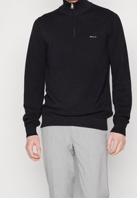 Zwarte gebreide pullover met een kwart rits en textuurstof. Voorzien van een klein logo op de borst. Gepaard met lichtgrijze broeken.