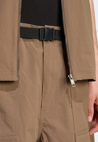 Kabat so silným opaskom v hnedej tkanine s hladkým povrchom, vybavený zipsom a detailmi ako prešité línie a malá logo nášivka.