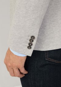 Ljusgrå kavaj i textil med struktur, fyra mörka knappar på ärmen och en ljusblå skjorta som tittar fram från under. Mörka denimjeans.