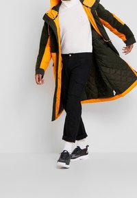 Orange och mörkgrön lång jacka med kviltad textur, parat med svarta cargobyxor och grå sneakers. Vit polo och korta strumpor synliga.