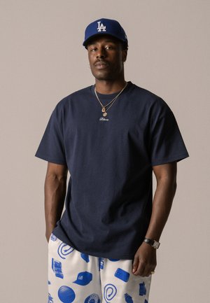 Navy t-shirt with short sleeves and a round neckline, paired with cream shorts featuring blue graphic prints. Accessories include layered necklaces.