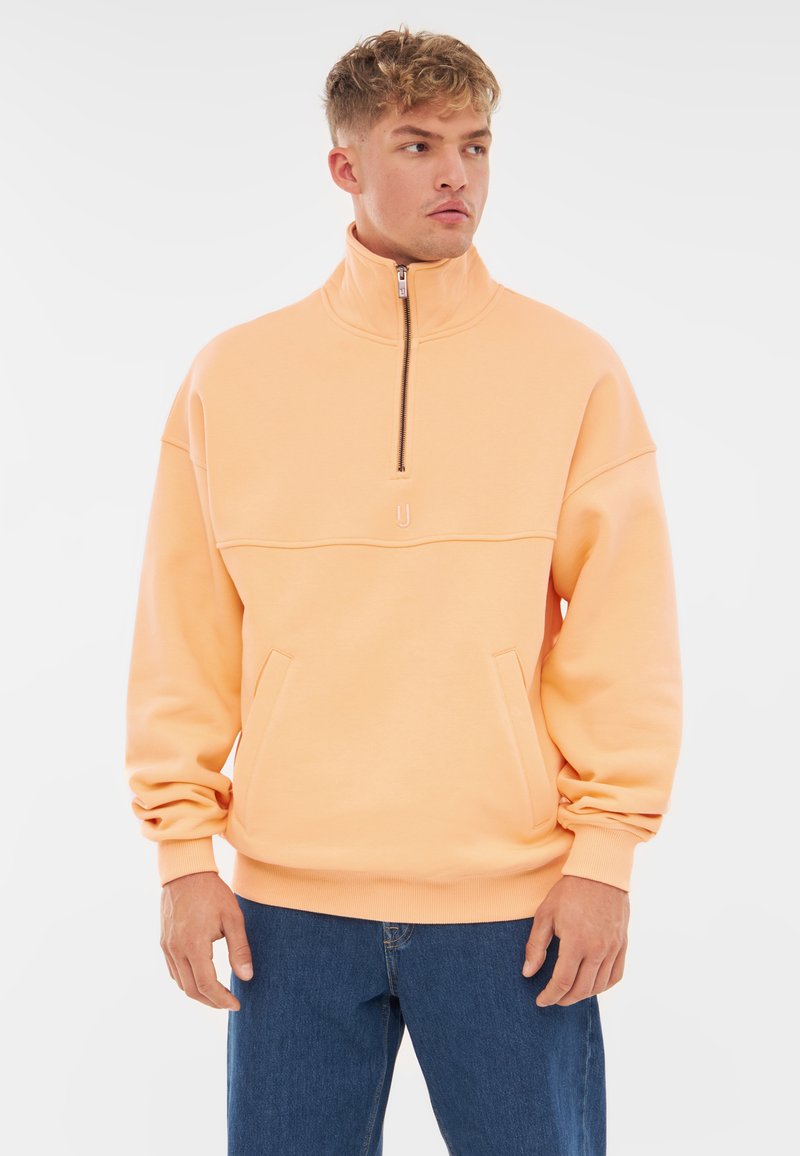 Oranje pullover met een hoge kraag, halve rits en voorzakken. Heeft een gladde textuur en ribbels aan de mouwen en onderkant. Groot formaat en ontspannen pasvorm.