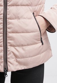 Person som bär en ljusrosa vadderad jacka med svart dragkedja, handen inskjuten i det dragkedjade sidofacket.