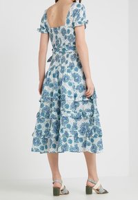 Blommig klänning med korta puffärmar, fyrkantig rygg, lager på lager volangkant, blå och grön mönster på vit tyg, tillsammans med klumpiga klacksandaler.