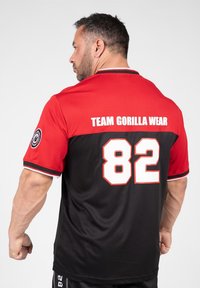 Korte mouw trui in rood-zwart stof, met de tekst "TEAM GORILLA WEAR" en het nummer "82" op de achterkant, voorzien van contrasterende randdetails.