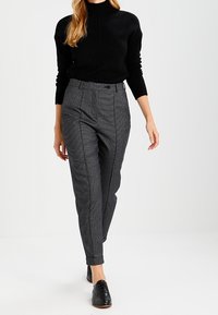 Schwarzer Strickpullover, kombiniert mit grauen Hosen im Karomuster. Die Hose hat eine hohe Taille und schmal zulaufende Beine, mit aufgerollten Säumen und vorderen Nähten.