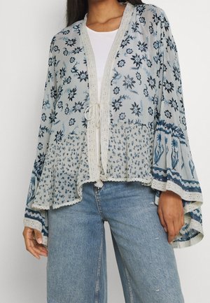 Vrouw draagt een los vallend kimono-achtig bovenstuk met een bloemmotief en brede mouwen, gecombineerd met lichtblauwe spijkerbroek en een wit shirt eronder.