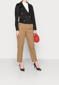Veste biker en daim noir avec fermetures éclair argentées, pantalon fuselé beige, talons noirs pointus et pochette rouge. T-shirt blanc simple en dessous.