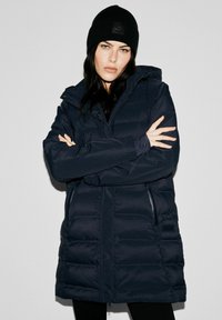 Granatowa kurtka puchowa z zamkiem z przodu, kapturem i bocznymi kieszeniami. Teksturowana tkanina o lekko połyskującym wykończeniu. Połączona z czarną dzianinową czapką beanie.