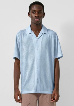 Jonge man die een lichtblauw overhemd met korte mouwen draagt, voorzien van verticale, getextureerde strepen, staand tegen een effen achtergrond.
