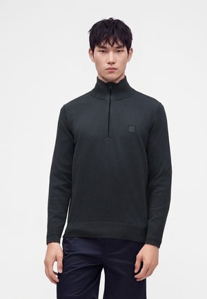 Junger Mann trägt einen dunkelgrauen Reißverschluss-Pullover mit hohem Kragen und dunkle Hosen, steht vor einem schlichten weißen Hintergrund.